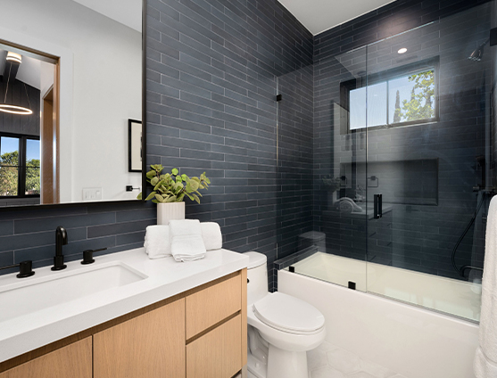 Modern bathroom with dark subway tile walls, glass shower enclosure, white countertop with sink, light wood vanity, black fixtures, and a decorative plant beside neatly folded white towels.
