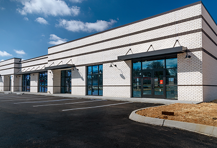 A modern, empty strip mall with white brick exterior, large glass windows, and vacant parking spaces under a blue sky with some clouds.
