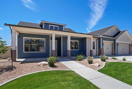 Single-story modern house with blue siding, large windows, a covered front porch with columns, and an attached three-car garage. The yard has green grass, landscaped shrubs, and a clear blue sky above.