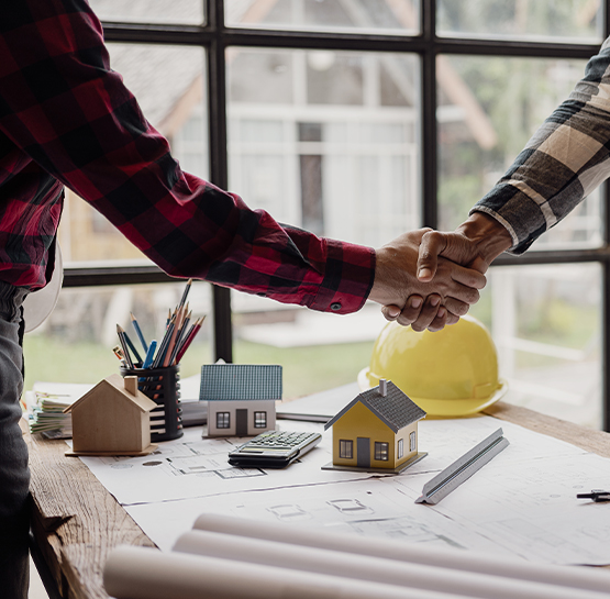Two people shake hands over a desk with house models, blueprints, a calculator, pencils, and a yellow hard hat, suggesting a real estate or construction agreement.