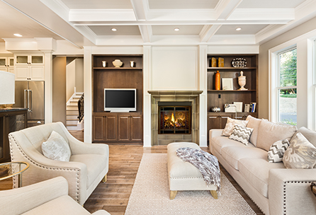 Bright, modern living room with beige sofas, armchairs, a fireplace, built-in shelves, a TV, and large windows letting in natural light. The room features hardwood floors and a coffered ceiling.