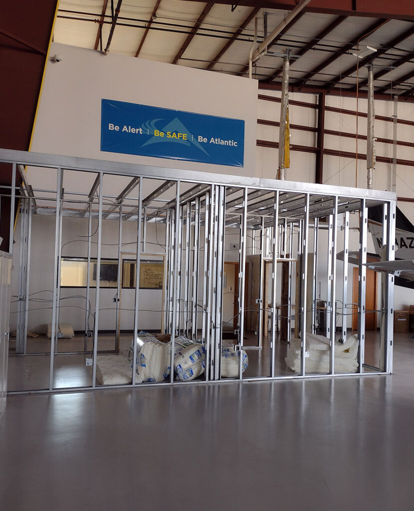 A partially constructed room with metal wall framing inside a large industrial building. Insulation rolls are on the floor, and a blue safety banner hangs above, reading “Be Alert | Be SAFE | Be Atlantic.”.