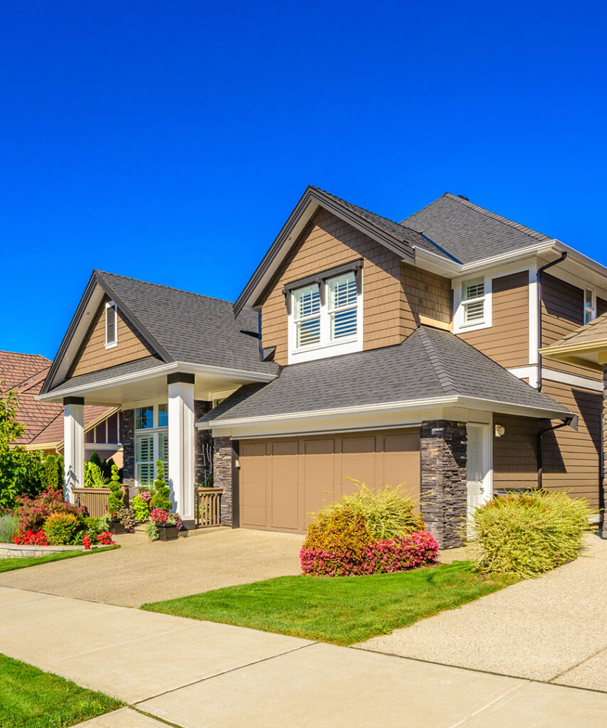 Modern two-story suburban house with brown siding, white trim, and a double garage, surrounded by green lawn, shrubs, and colorful flowers under a clear blue sky.