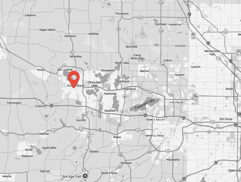 A grayscale map highlighting Oconomowoc, Wisconsin with a red location pin. Surrounding towns and cities, roads, and shaded areas indicating geographic features are also visible.
