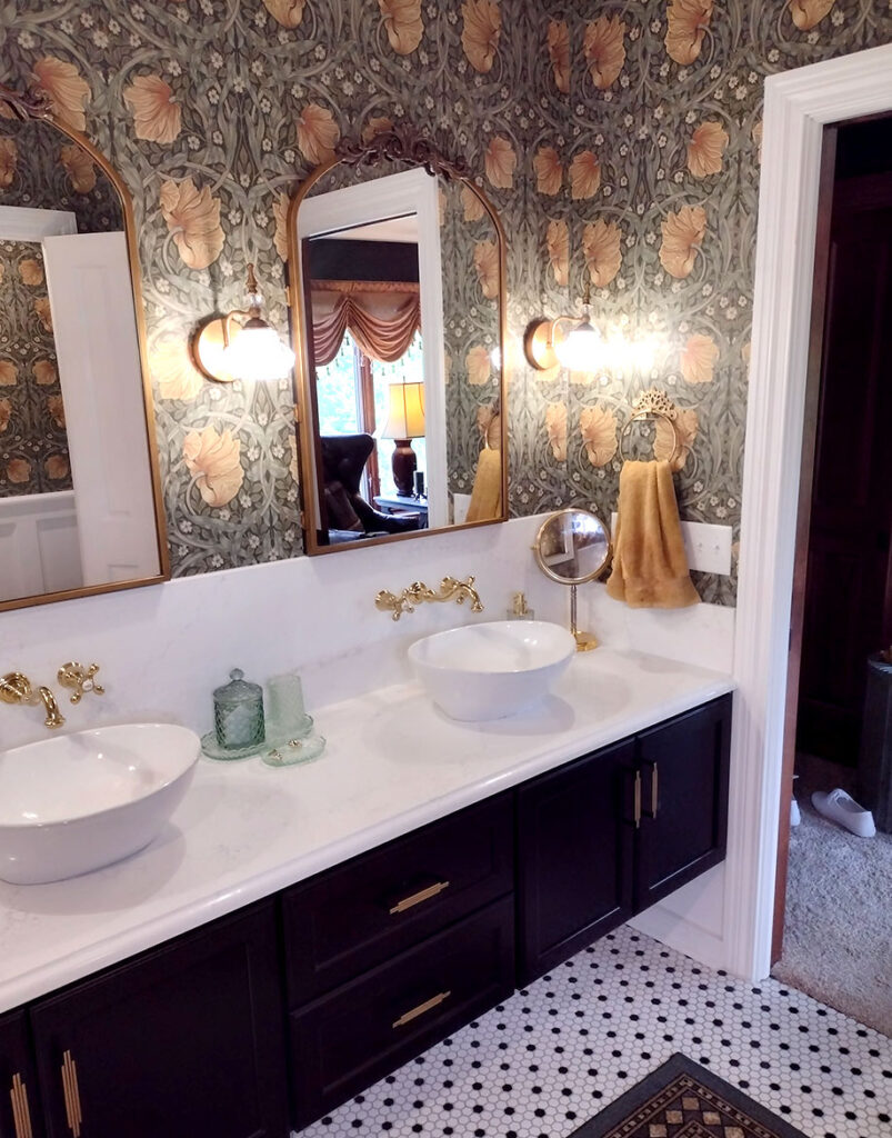 A bathroom with dark cabinets, two vessel sinks with gold fixtures, a white countertop, floral-patterned wallpaper, two mirrors, wall sconces, and towels hanging on the wall. The floor has a tile pattern.